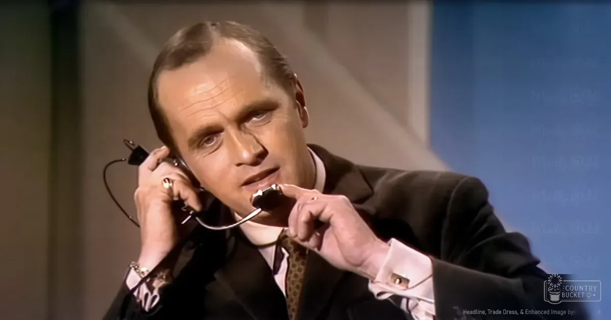 A middle-aged man wearing a dark suit jacket with light-colored cuffs and lapels, white dress shirt, patterned tie, and wristwatch, holding a telephone to his ear in an indoor setting.