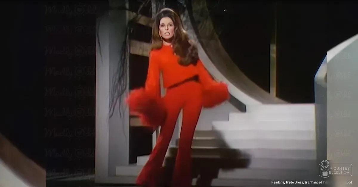A woman wearing a bright orange bell-bottom jumpsuit with a high neckline stands outdoors at night under artificial lighting.