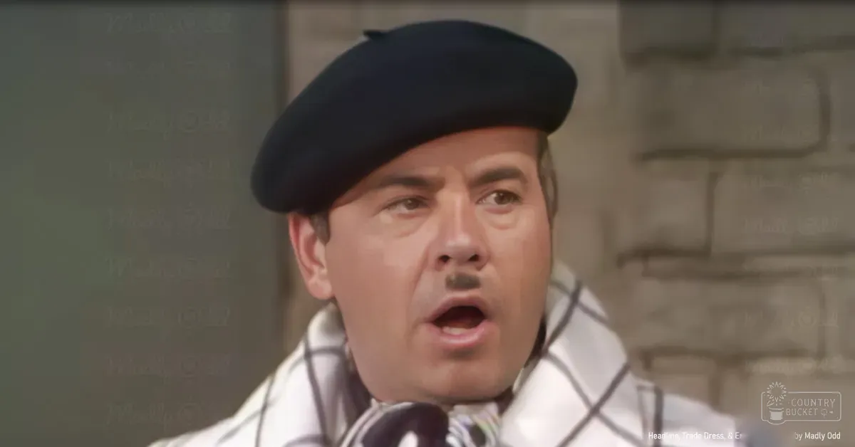 A middle-aged adult male wearing a black beret-style hat and a white garment with black checkered patterns, set against an indoor brick wall background during daylight.