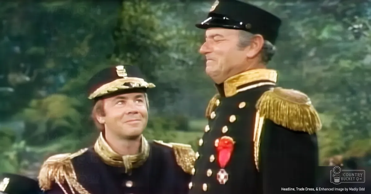 Two men in historical military uniforms stand outdoors with greenery and natural light, suggesting a spring or summer setting.