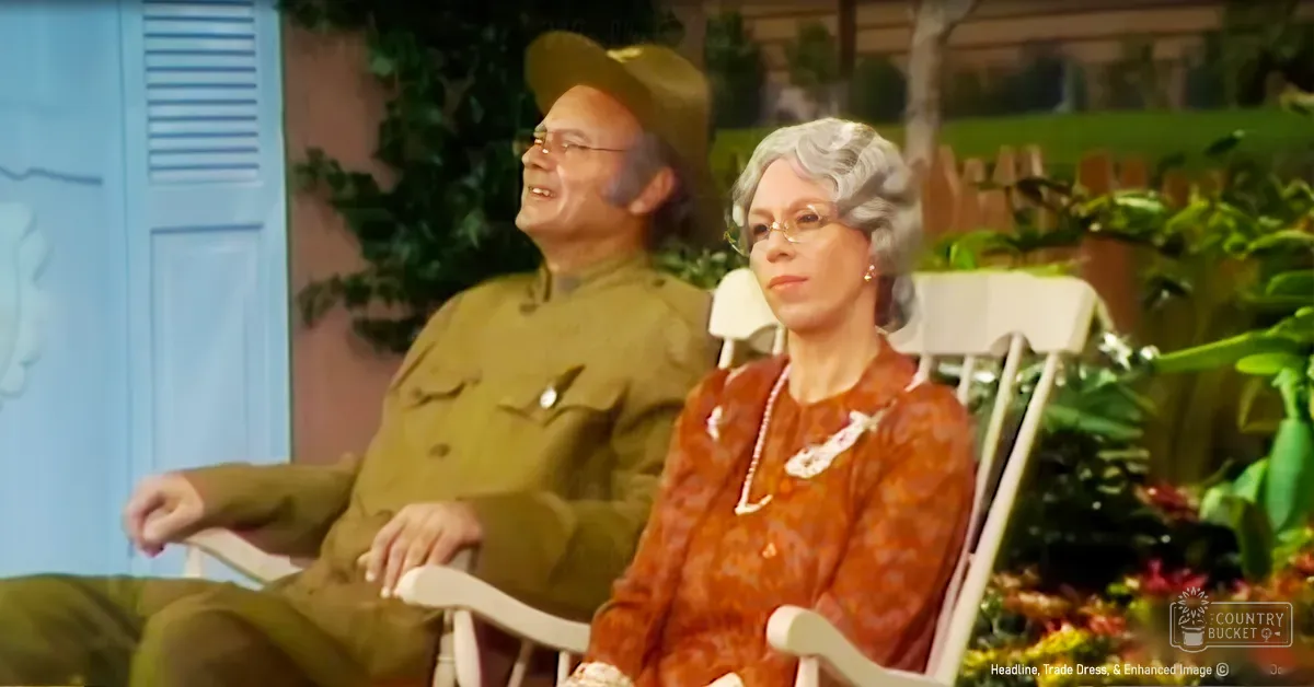 An older adult couple in a lush garden setting with greenery and white plastic chairs, wearing a military-style jacket and an orange floral dress under natural daylight.