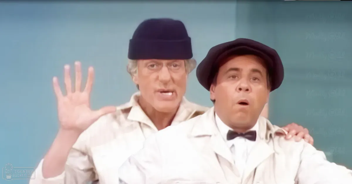 Two middle-aged men in a classroom setting with a chalkboard and light blue walls; one wearing a beanie, glasses, white shirt, and jacket; the other in a dark cap, bow tie, white shirt, and loose-fit coat.