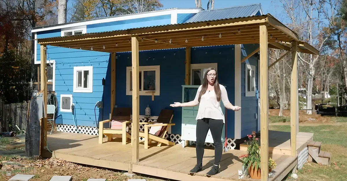 An adult female stands outdoors, wearing a loose-fitting white blouse with long sleeves and black leggings and ankle boots, surrounded by a small blue house with wooden pergola furniture and string lights in a residential backyard setting during late autumn or early spring.