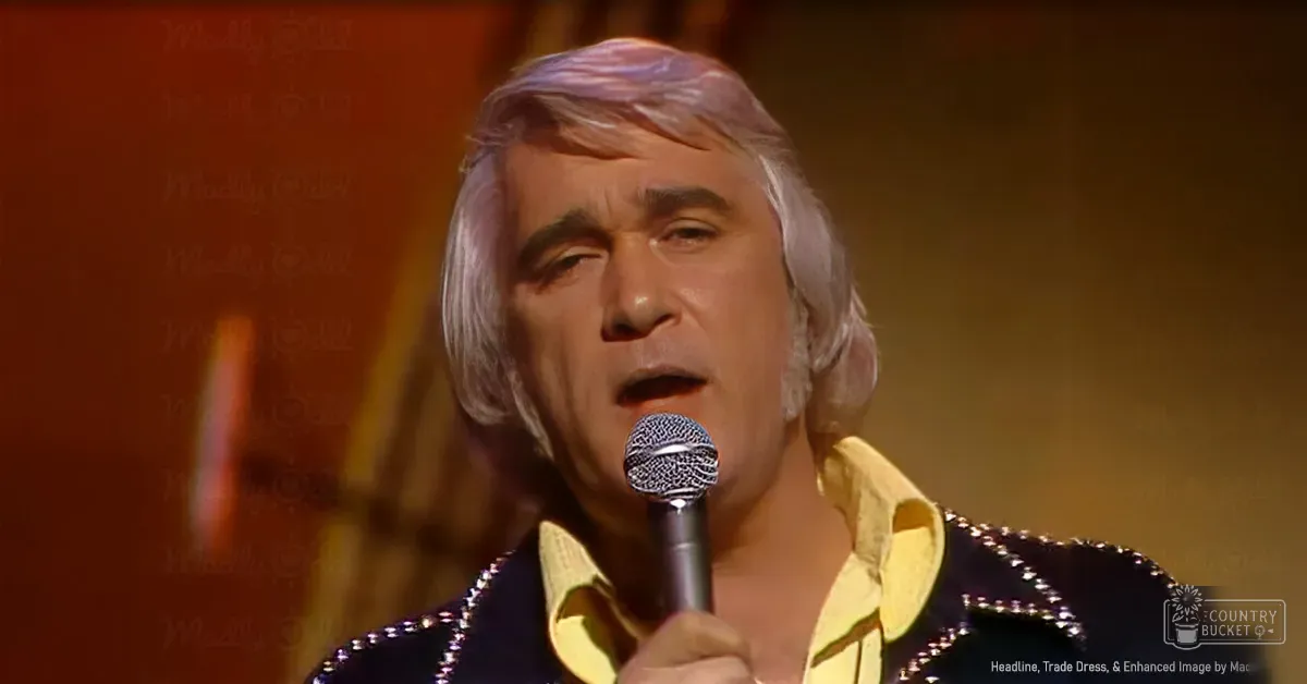 A middle-aged to older male performer wearing a yellow shirt with black sequined or beaded jacket, holding a microphone in an indoor venue with warm lighting.