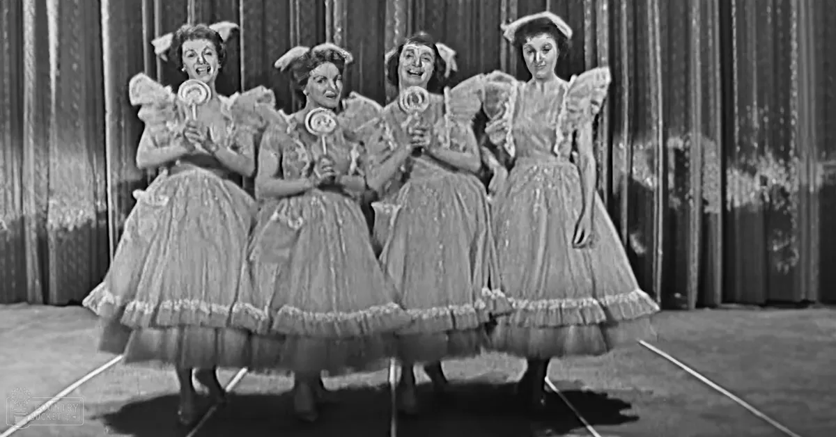 Group of performers on stage holding lollipops; theatrical setting with dark curtain backdrop and indoor lighting.