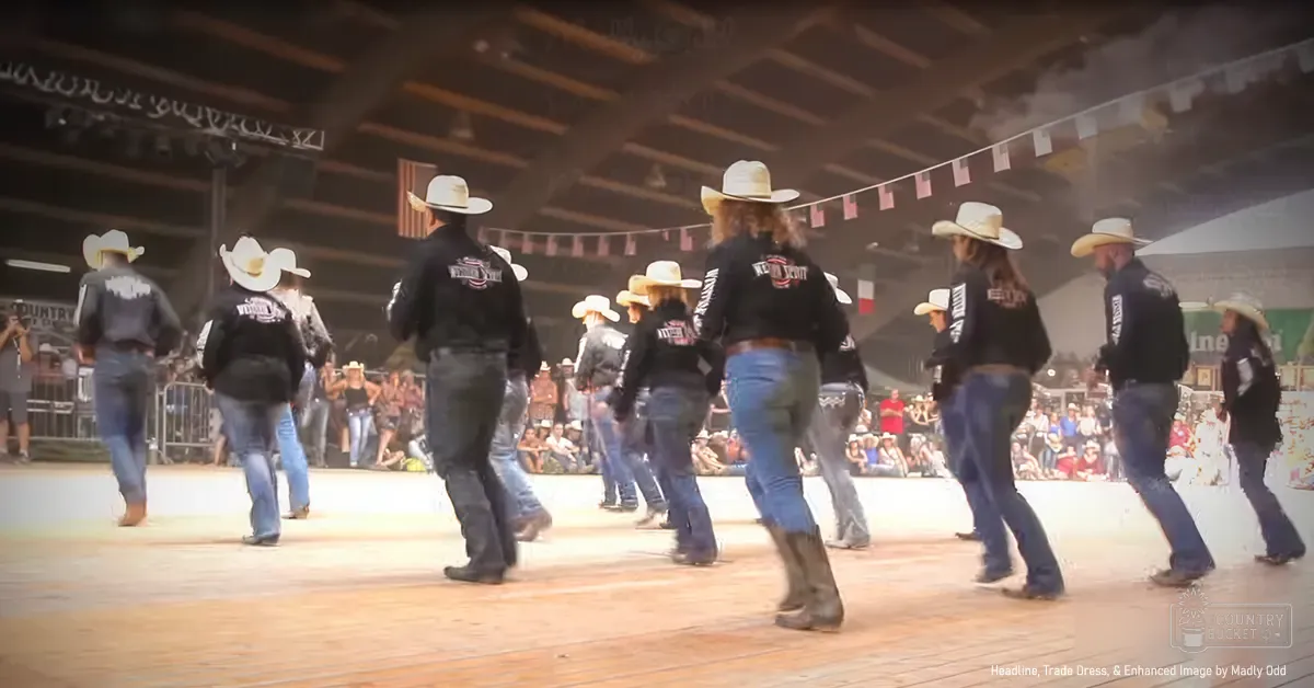 Young adults performing a dance routine at an indoor rodeo or country music event with spectators seated on bleachers, wooden floor, and artificial lighting.