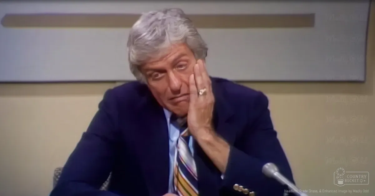 Older adult male in formal attire, dark blue suit jacket with gold buttons and light blue dress shirt with yellow, red, and white striped tie, microphone partially visible, indoor setting possibly conference room or meeting space.