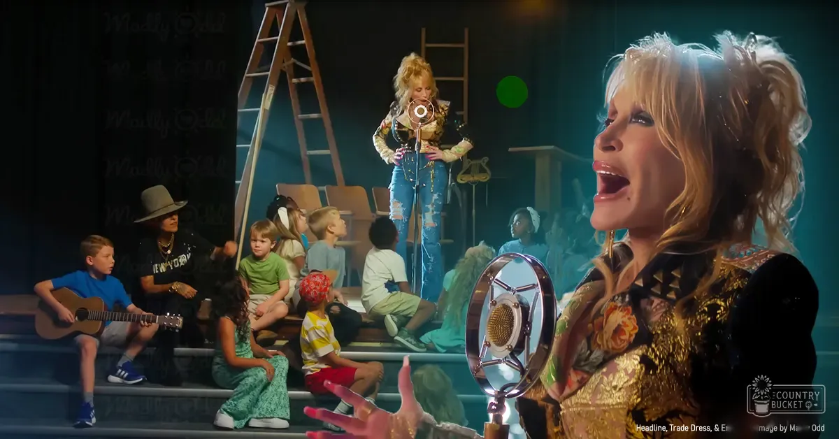 A performance scene with a female performer wearing a black and gold patterned top and large hoop earrings singing into a microphone while another performer on a ladder holds the stand, surrounded by children in casual clothing against a dark backdrop.