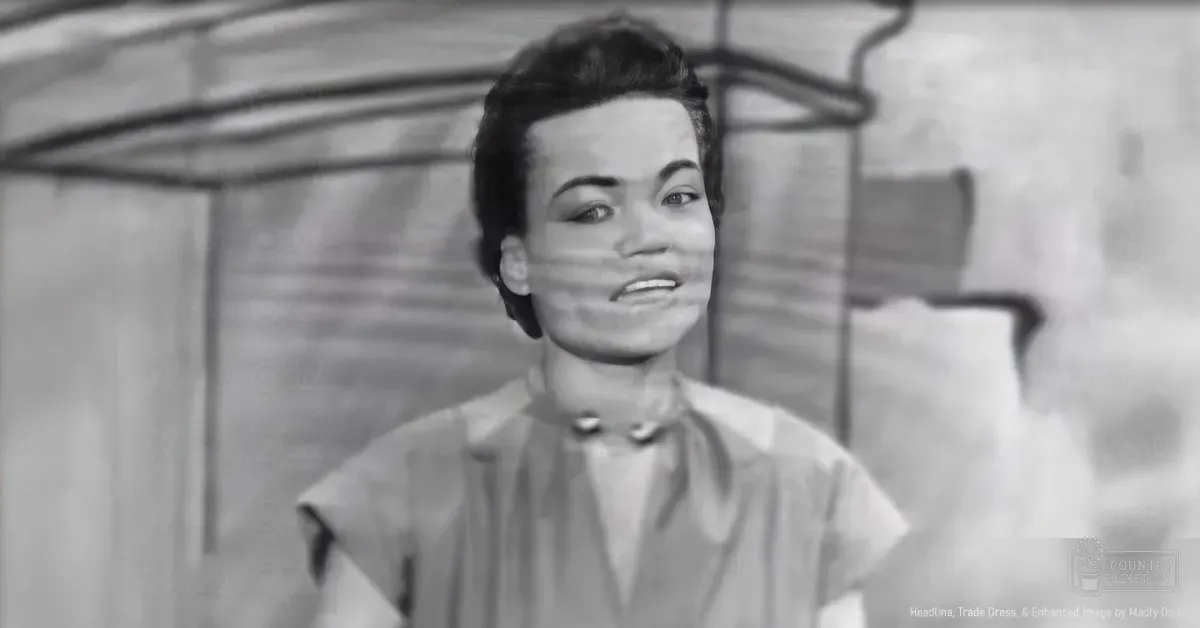 A young adult woman wearing a tailored blouse indoors with an industrial brick wall backdrop.