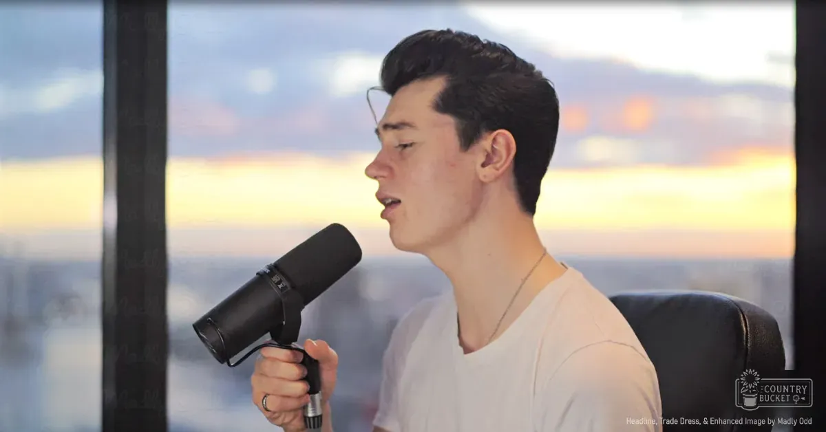 A young adult male in a relaxed fit white t-shirt stands indoors, with a cityscape view through large windows during sunset or sunrise.