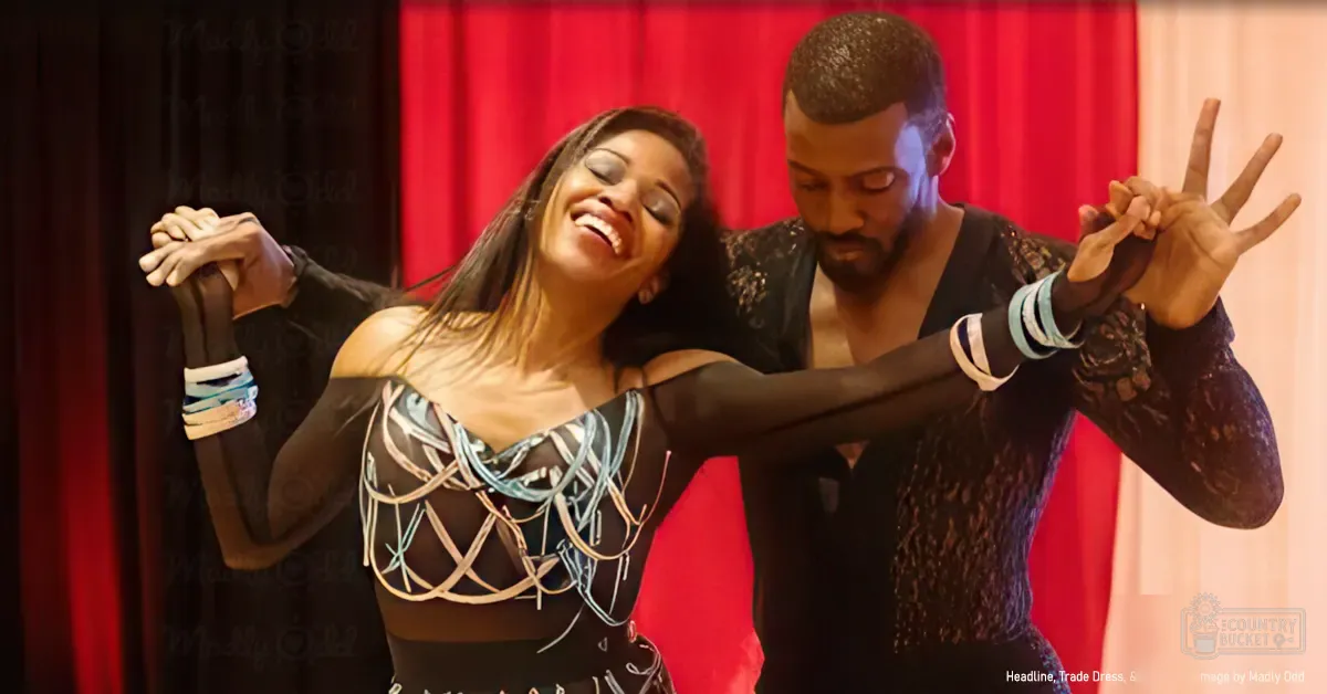 A dance performance scene with two dancers, one in a black and white patterned top and another in a black lace-like top, set against red curtains indoors under bright lighting.