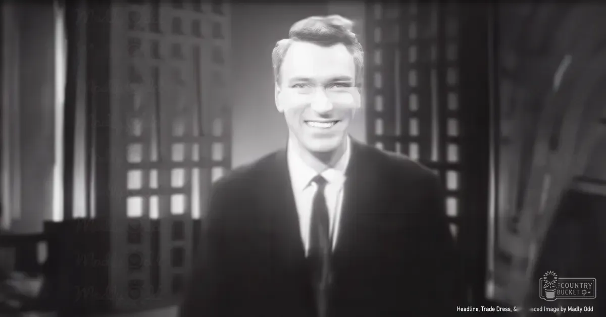 Young adult male in a dark suit jacket with a classic mid-20th century cut, standing indoors near large windows or doors during daytime.