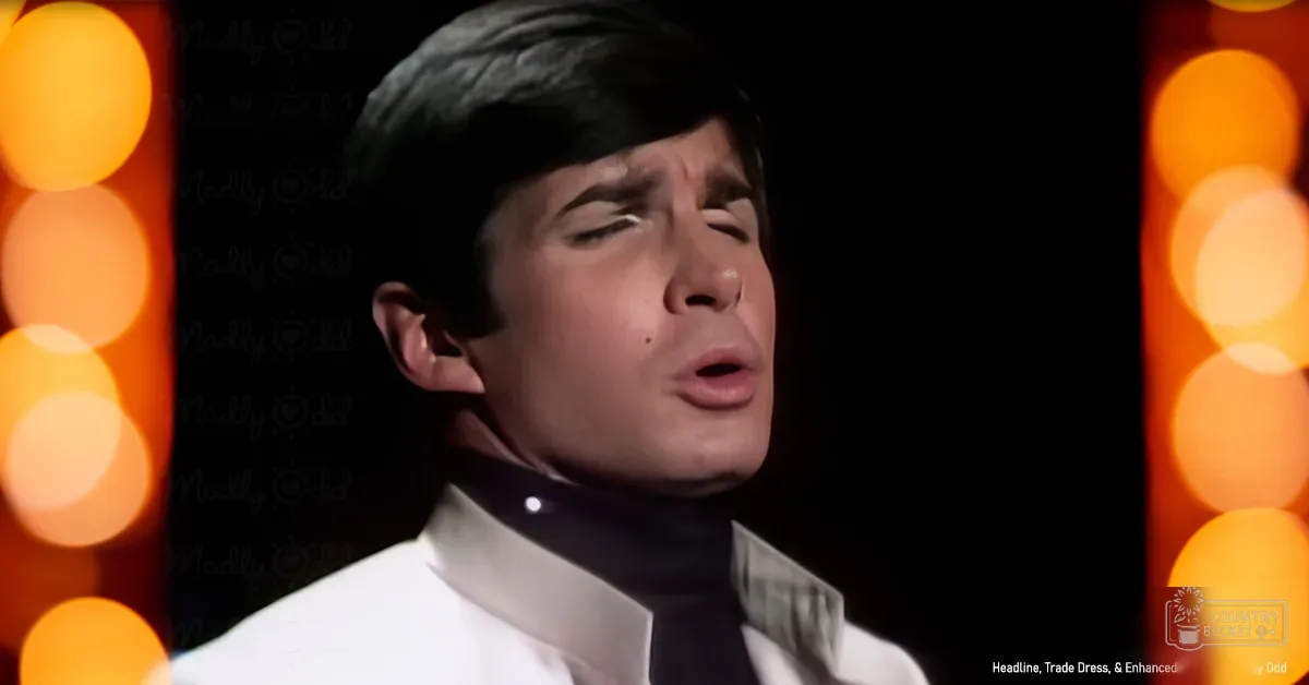 A young adult male in a white dress shirt and black tie stands against an orange-yellow bokeh-lit backdrop, suggesting a stage-like setting.