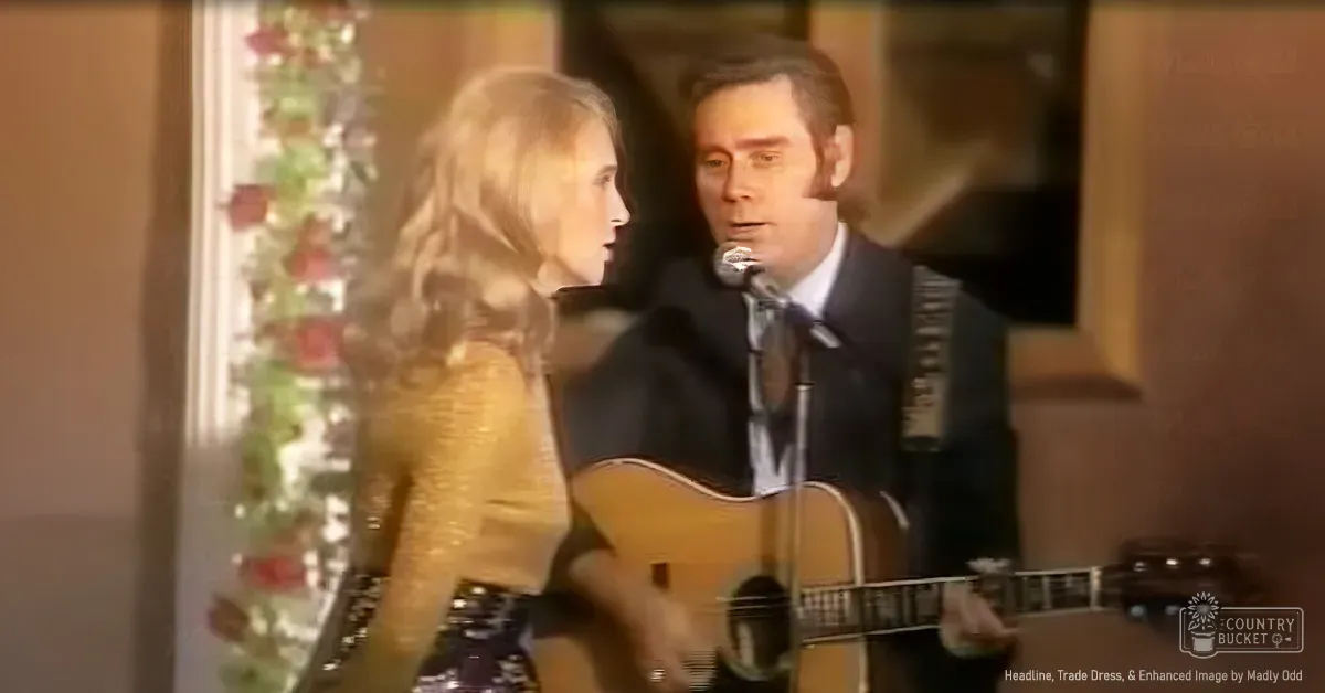 Musical performance with an adult female in a patterned top and middle-aged male holding an acoustic guitar, set indoors with decorative garlands and warm lighting.