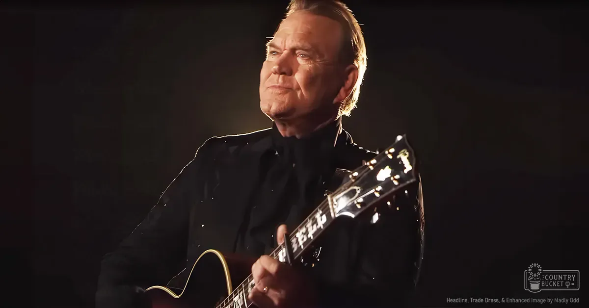 A middle-aged man playing an acoustic guitar in a dimly lit studio setting with dramatic lighting.