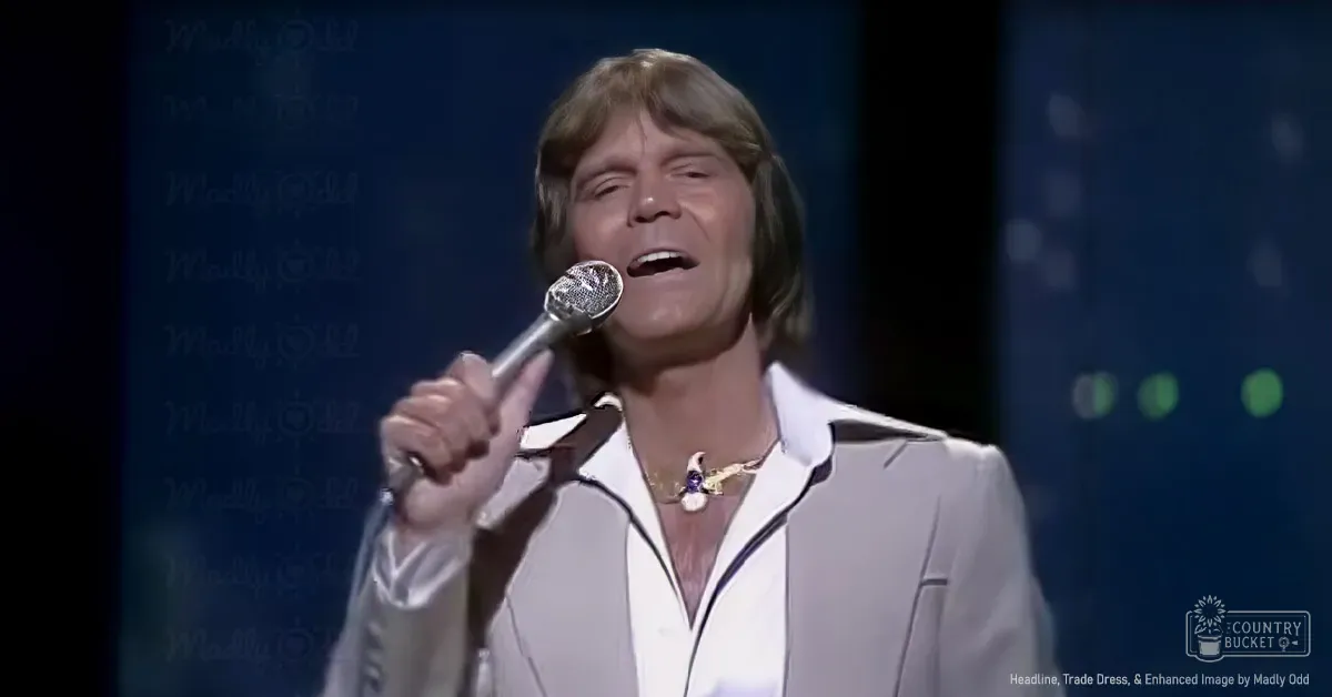 A middle-aged male performer singing into a microphone wearing a white jacket with black trim in an indoor studio or stage setting.