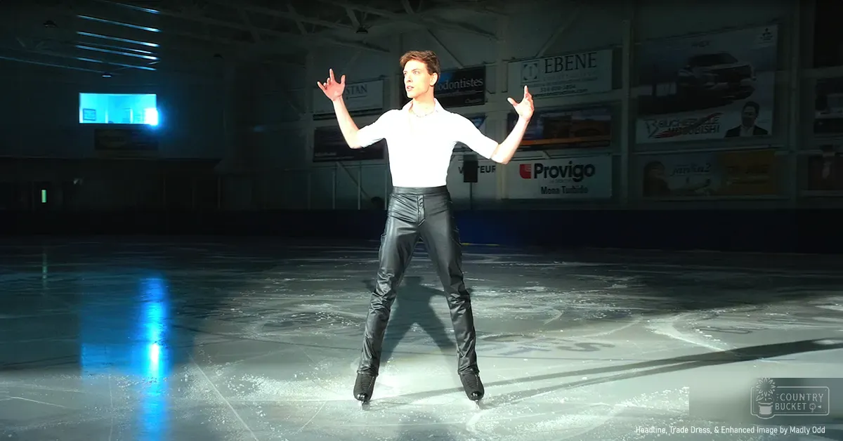 A young adult male figure skater in a modern era white long-sleeved shirt, black leather pants, and figure skate boots stands at an indoor ice rink with advertisements for businesses like EBENE visible on the walls.
