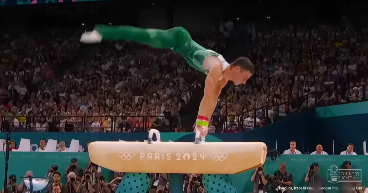A male gymnast performs on a light-colored pommel horse at an indoor arena during the Paris 2024 Olympics, with spectators in casual attire and evening stadium lights illuminating the scene.