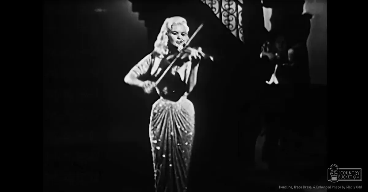 A female adult performer wearing a long, embellished dress with a fitted bodice and flared skirt, playing the violin on what appears to be a stage set in an indoor venue.