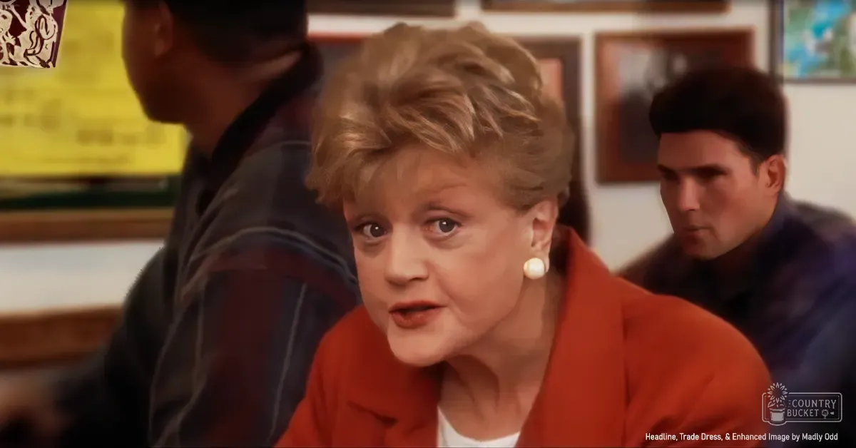 A middle-aged woman wearing a red blazer over a white top stands indoors with a young adult male in the background, both dressed from the 1980s to early 1990s. A yellow sign and framed artwork are visible behind them.