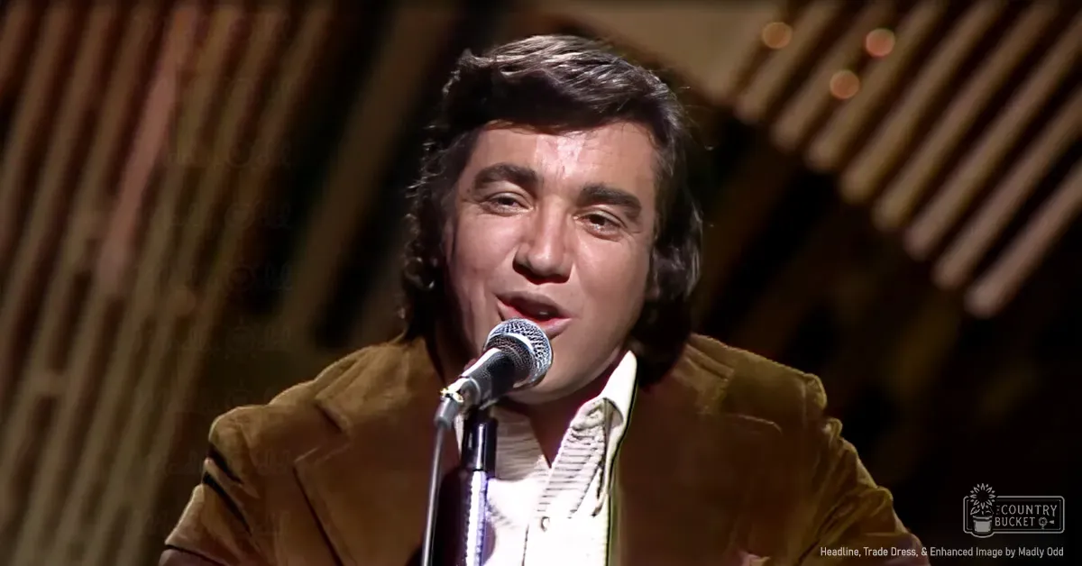 Young adult male wearing a brown jacket over a white button-down shirt with vertical stripes, performing on stage with a microphone and wooden paneling in the background.