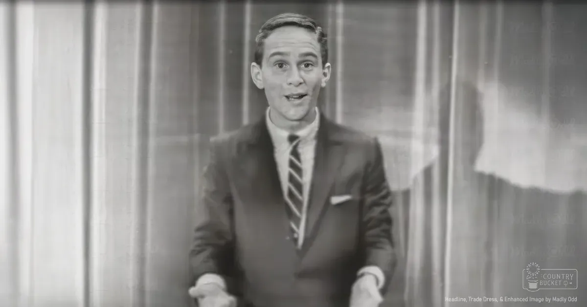 A young adult male wearing a dark suit jacket with a white pocket square, standing indoors against vertical blinds or curtains.