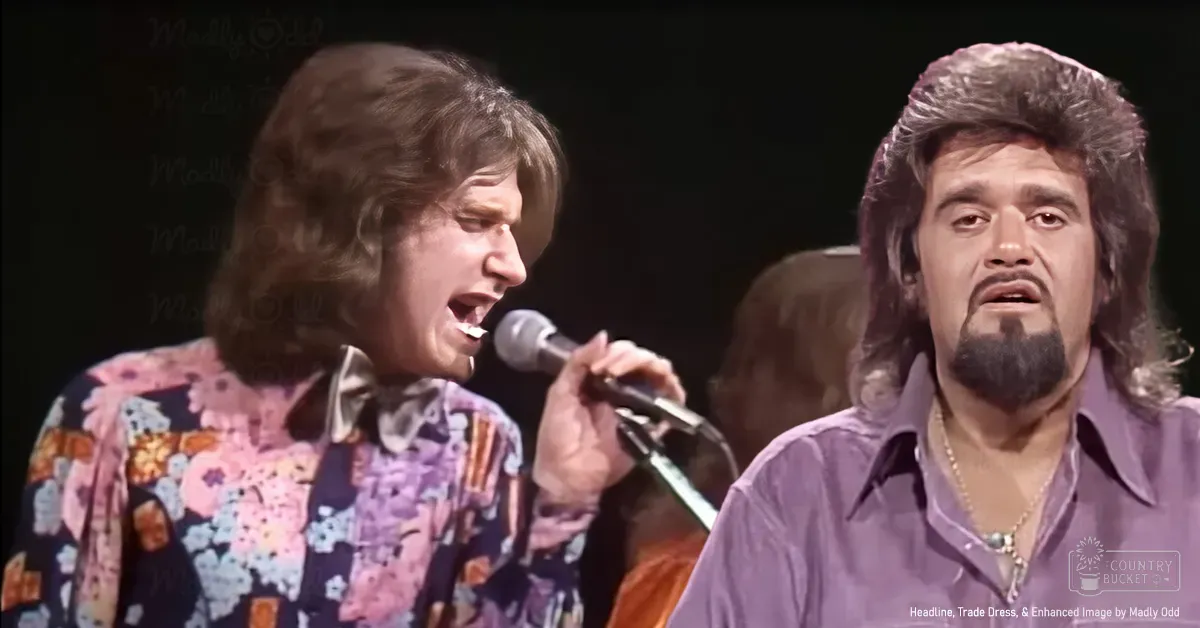 A musical performance scene with a young adult male singer in a floral-patterned shirt and an older male performer wearing a purple button-down shirt with a gold chain, set on a stage with dark curtains under spotlighting.
