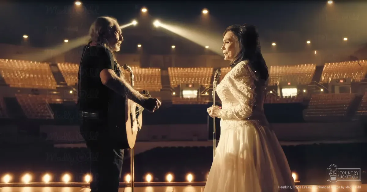 An older adult male singer wearing a sleeveless black shirt with tattoos performing in an indoor arena or stadium alongside another older adult female, both dressed for a musical performance under stage lighting during what appears to be the evening.