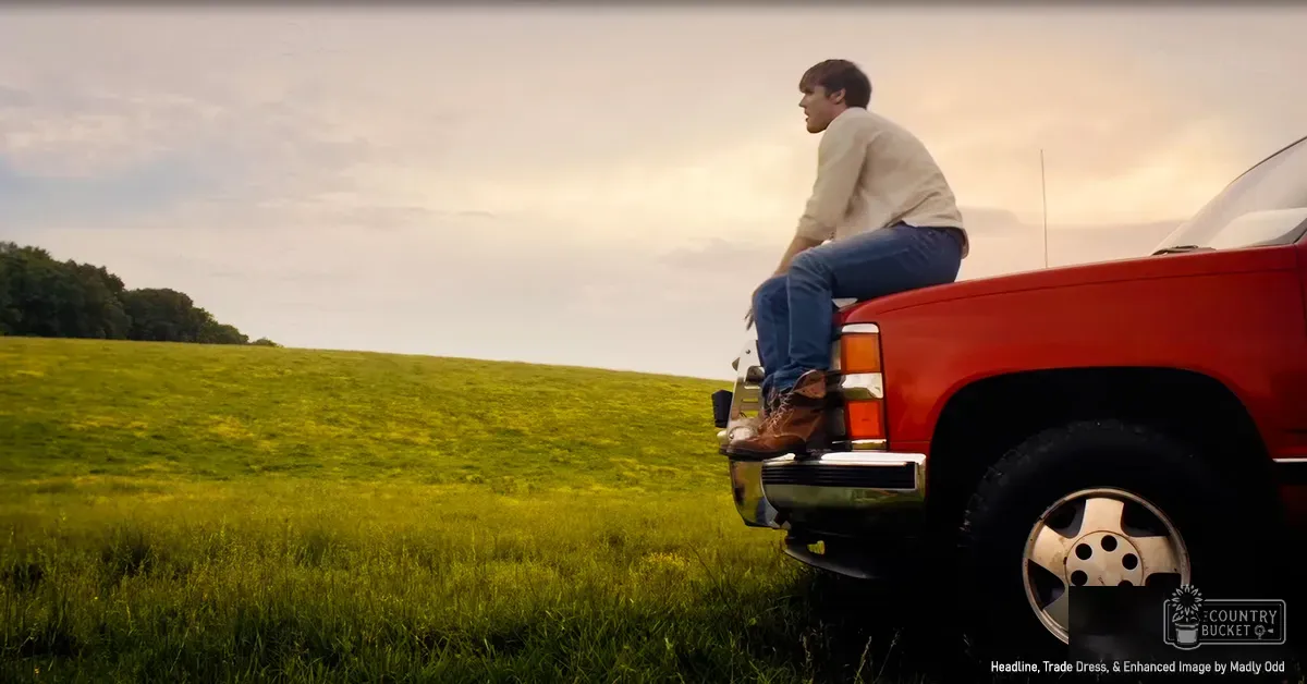 A young adult male in a relaxed outfit sits on the tailgate of an older red pickup truck in an open field with green grass and trees under a cloudy sky, suggesting late afternoon or early evening light.