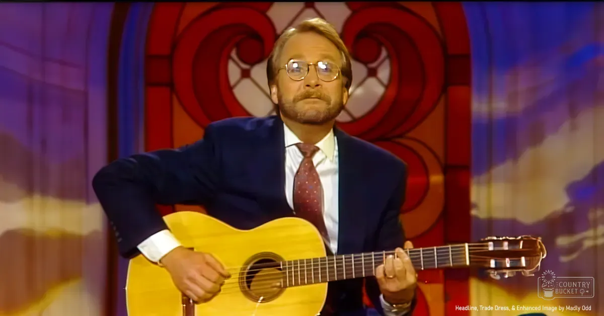 A middle-aged adult male in a formal dark blue suit with a red dotted tie holds an acoustic guitar on stage, set against a colorful stained glass backdrop.