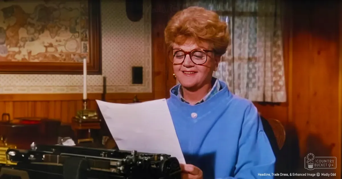 An older adult woman wearing a blue smock with button detail in an indoor setting, vintage typewriter on the table.