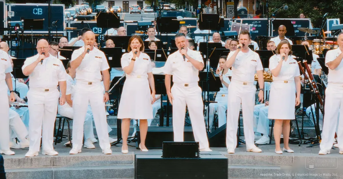 Adult males in white dress uniforms with gold belts perform on an outdoor stage surrounded by urban scenery during daytime, likely spring or summer.