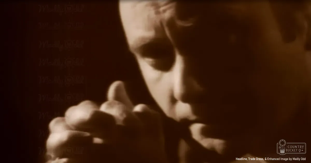 A young adult male dressed formally in a dark-colored suit with a bow tie, holding a small object near his face against a sepia-toned indoor backdrop.