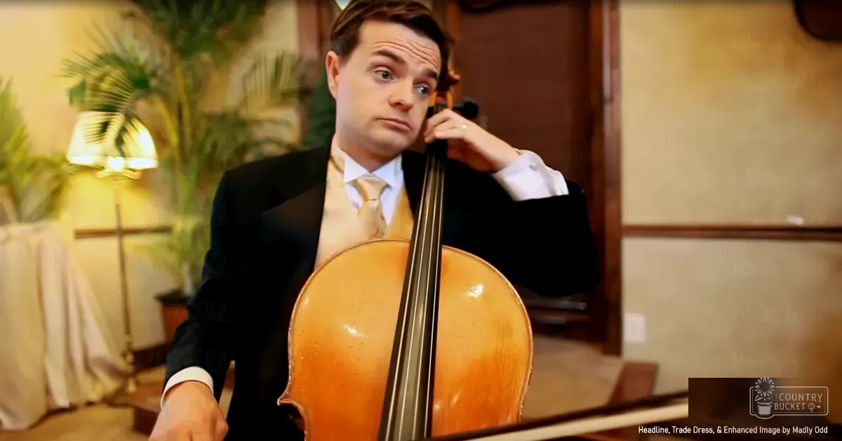 An adult male playing a cello indoors with warm lighting, surrounded by plants and textured walls.