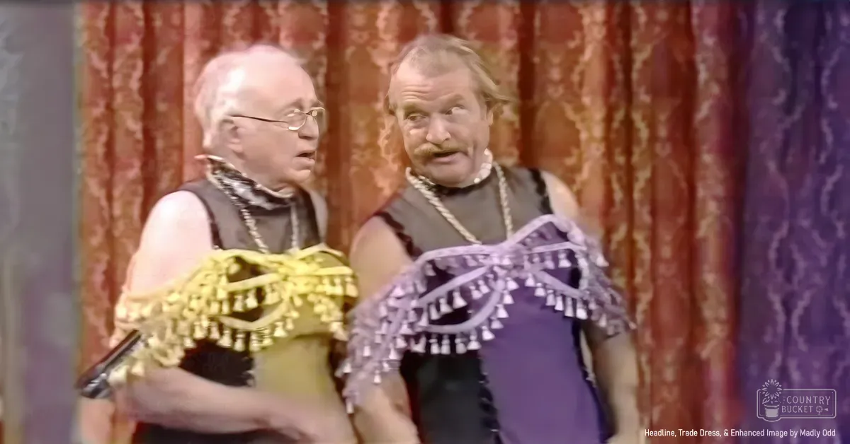 A comedic scene with two older adult males wearing loose-fitting sleeveless garments inspired by traditional Native American attire, holding a microphone and adorned with beads.
