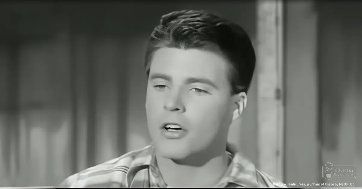 Young adult male wearing a light, collar-closed shirt in an indoor setting with wooden paneling.