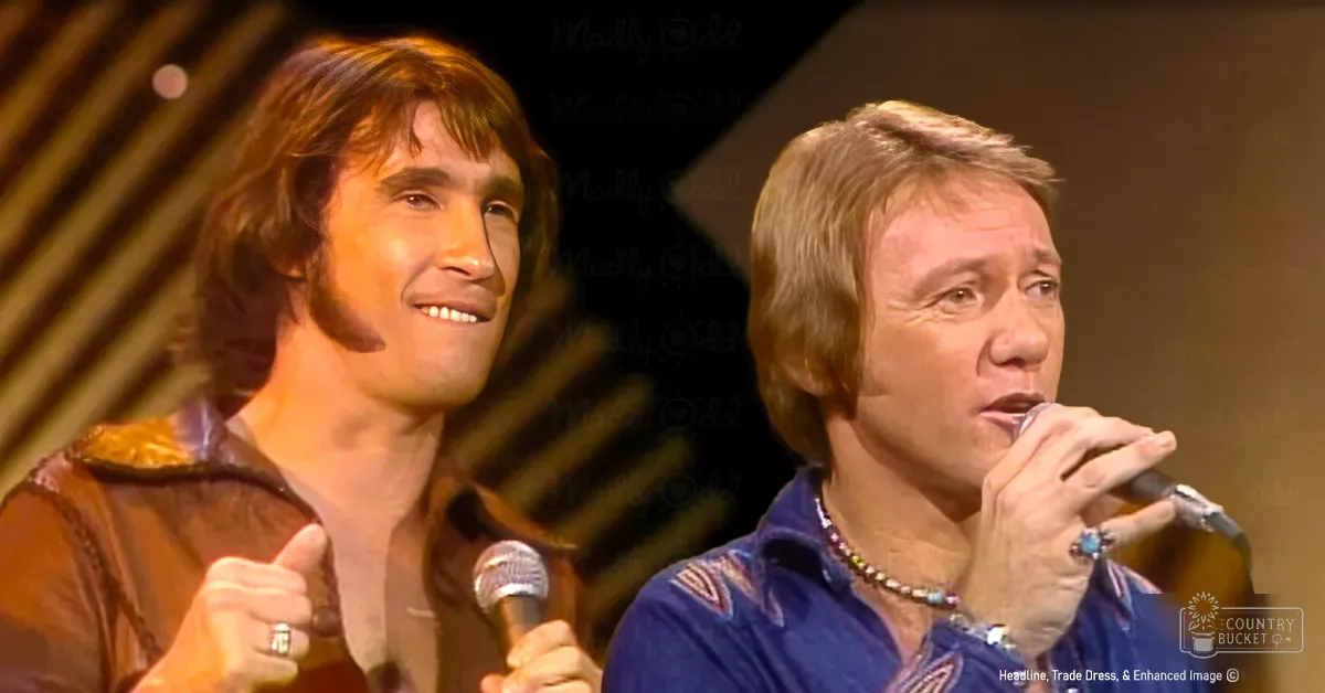 Two adult male performers on a concert stage, one wearing a brown fur-trimmed jacket and another in a blue striped shirt; each holding microphones and accessorized with rings and necklaces under warm stage lighting.