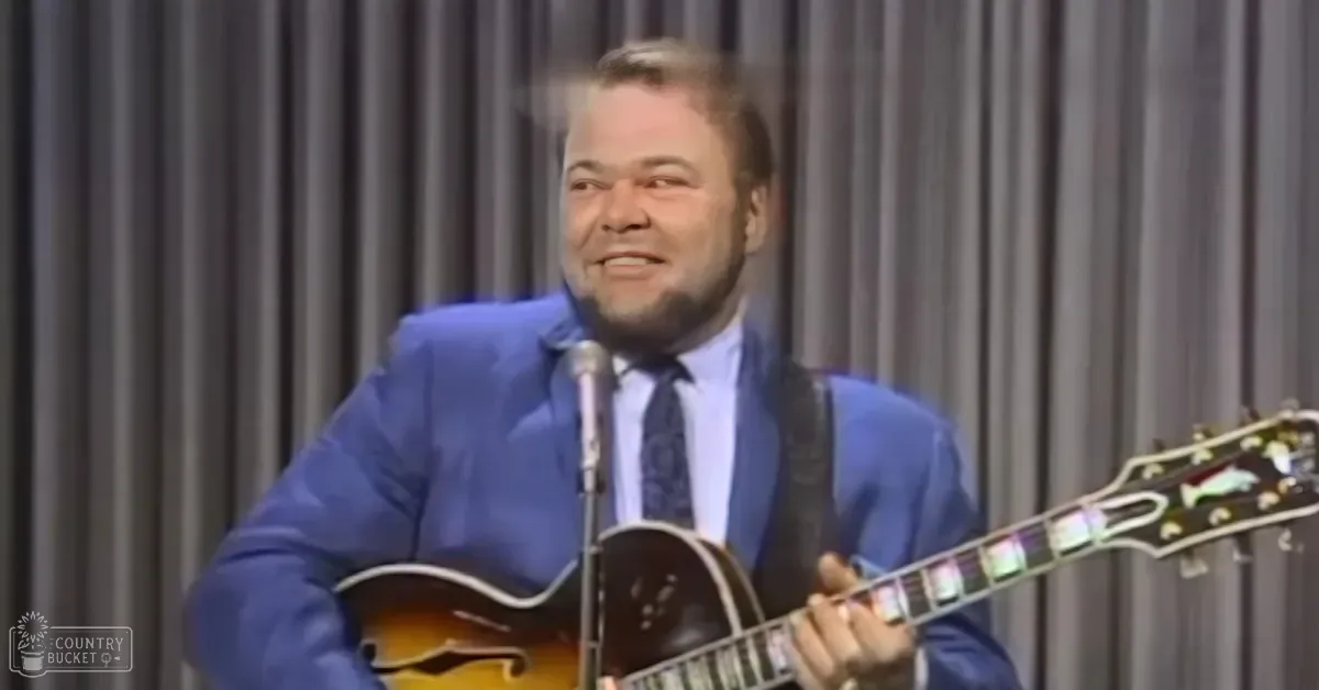 Adult male performer in a blue suit jacket and white shirt, holding a guitar with sunburst finish on stage. Indoor setting with gray curtains as backdrop.