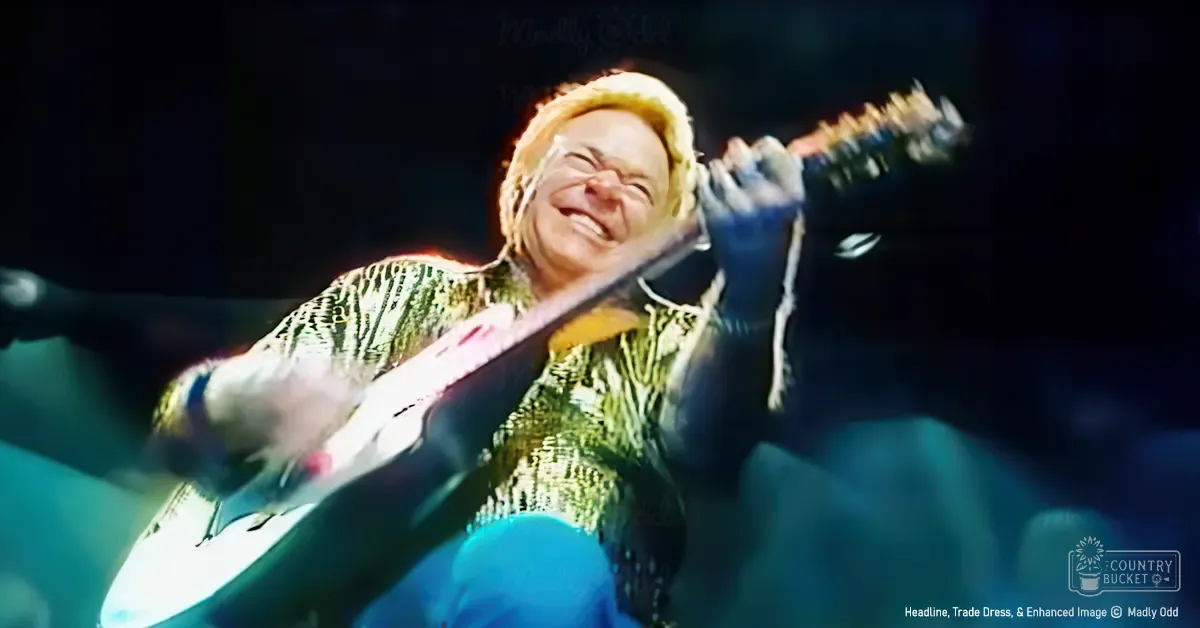 A middle-aged male singer wearing a shiny, sequined 1970s-style shirt performs at an indoor concert arena with stage lights and audience in the background.