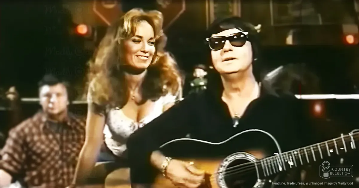 A middle-aged man plays a guitar, wearing sunglasses and a black shirt with a wristwatch, while an adult female in a white top and necklace stands in the background; stage lighting illuminates an indoor venue.