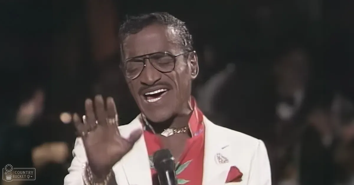 A middle-aged adult male performer in a white suit jacket with floral lapels, red scarf, and glasses, holding a microphone on stage in an indoor concert hall setting.