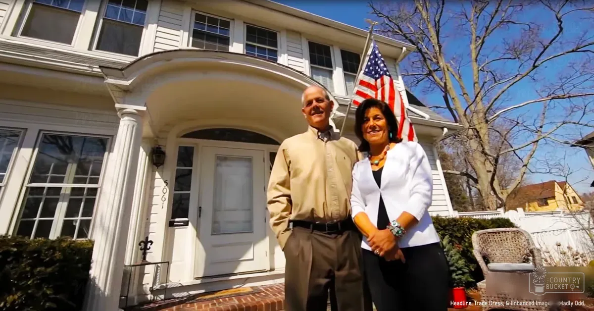 A couple stands in front of their suburban house on a clear day with bare trees suggesting late autumn or early spring. The scene includes white siding, brick foundation, an American flag, and a wicker chair.
