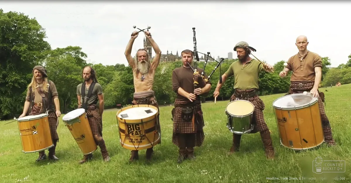 Adult males performing traditional Scottish music in a park with greenery, wearing plaid skirts and tunics, holding bagpipes and drums under an overcast sky.