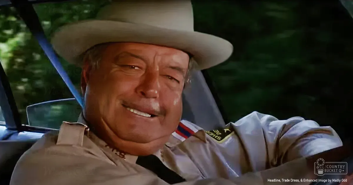 A middle-aged adult male in a vehicle, wearing a light beige uniform shirt with epaulets and a wide-brimmed hat, driving during daytime in spring or summer, with green foliage visible through the windows.