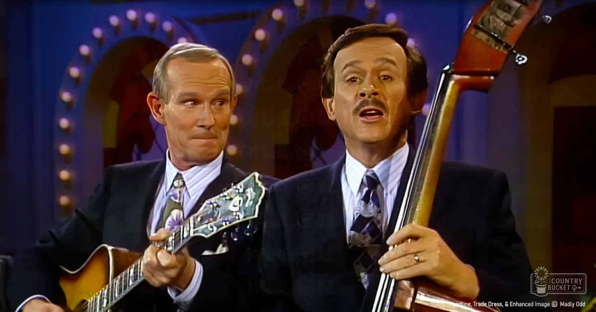 A musical performance scene with two middle-aged male musicians, one playing a guitar and the other holding a double bass, set against an indoor stage backdrop with decorative arches and lights.