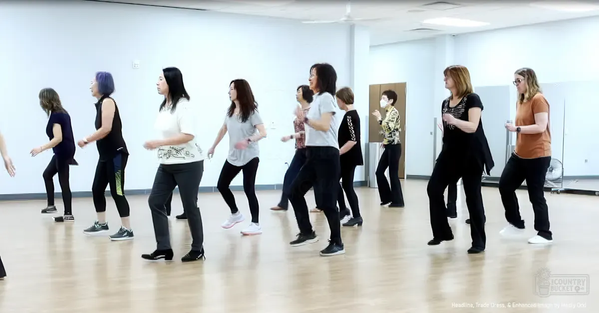 A group dance class in an indoor studio with light-colored walls, wooden flooring, and a whiteboard at the back. Light beige floor, white walls, black clothing items, gray pants, orange t-shirt, various patterns on tops.