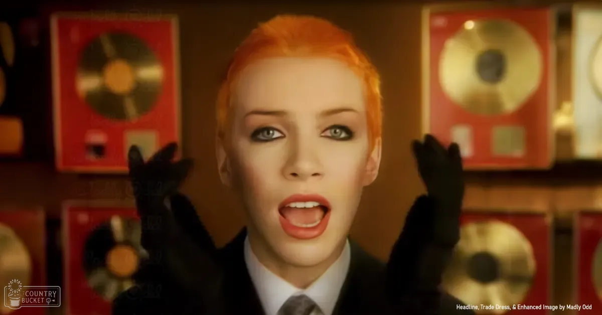 A young adult female in a formal suit with a patterned tie, standing indoors amidst framed awards related to the music or entertainment industry.