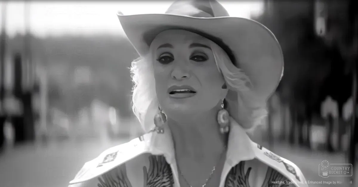 An older adult woman in an outdoor setting at a fair or event wearing a wide-brimmed white hat, a white shirt with decorative patches on the shoulders and chest area, large hoop earrings.