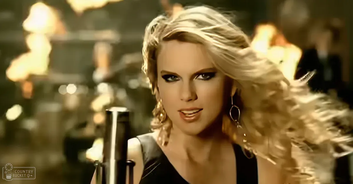 A young adult female in a modern pop music style, wearing a black sleeveless V-neck top with large hoop earrings and holding a microphone stand in an indoor music performance space. The setting is dimly lit with warm tones from background lights.