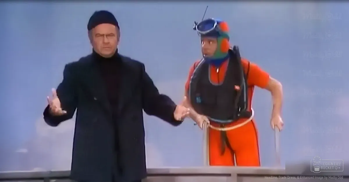 A middle-aged male wearing a dark coat stands indoors with another person dressed as an underwater diver, who is in an orange wetsuit and hooded design against a light blue backdrop.